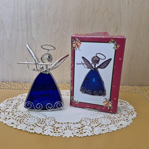 May include: A decorative angel figurine with a blue stained glass dress and silver wire accents. The angel has a halo and wings, and is next to a box with a similar image. The figurine is on a white lace doily.