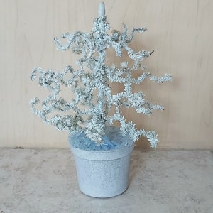 May include: A white, artificial Christmas tree made of curled paper, in a white pot with a blue base. The tree is decorated with white glitter.