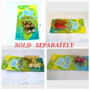 May include: A collection of small clay figurines, each packaged on a yellow and green card with the text "Bitty Buddies." The figurines include a butterfly, carrots, dogs, and ladybugs. The words "SOLD SEPARATELY" are also visible.