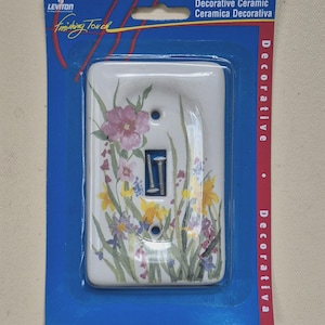 May include: Decorative ceramic light switch cover with a floral design. The white cover features a watercolor-style painting of pink, yellow, and purple flowers with green foliage. The cover is in a blue plastic package.