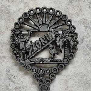 May include: A dark gray metal trivet with a circular design. The trivet features the word "FLORIDA" and images of a Native American, palm tree, alligator, and a bird. The trivet has a handle and a sunburst design.