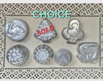 May include: A collection of vintage metal molds in various shapes, including a rabbit, a Christmas tree (marked "SOLD"), and playing card suits. The word "CHOICE" is above the molds. The molds are displayed on a decorative, lacy tray.