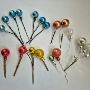 May include: A collection of vintage Christmas tree ornaments in various colors, including blue, red, gold, and silver. The ornaments are spherical and attached to stems, some with wire and others with wooden sticks.
