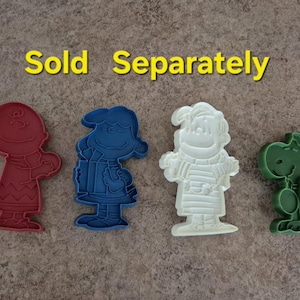 May include: Four colorful cookie cutters in the shapes of cartoon characters. The cutters are red, blue, white, and green. The text "Sold Separately" is in yellow above the cutters.