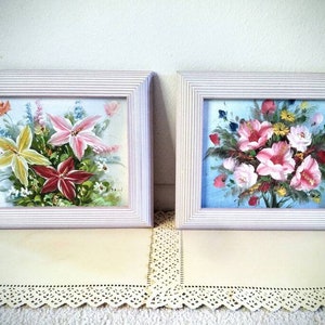 May include: Two framed floral paintings with white frames. The paintings feature colorful flowers in a vase. The paintings are on a white lace doily.