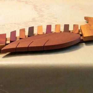 K&ouml;nnte beinhalten: Eine Holzskulptur eines Vogels mit einem segmentierten K&ouml;rper, der in Braun-, Rot- und Lilat&ouml;nen bemalt ist. Der Vogel hat einen langen, spitzen Schnabel und einen abgerundeten Schwanz.
