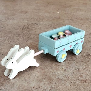 May include: A light blue wooden wagon with two white bunny pull toys. The wagon is filled with colorful candy eggs and has a floral and heart design.