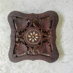 May include: A square, dark brown wooden trivet with intricate leaf and floral carvings. The center features a circular design with a white starburst pattern. The edges are scalloped, adding a decorative touch. A handcrafted item.