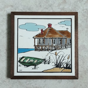 May include: A framed tile with a hand-painted illustration of a beach house with a brown roof, a green boat on the sand, and tall grass with brown seed pods. The tile is surrounded by a dark brown wooden frame.