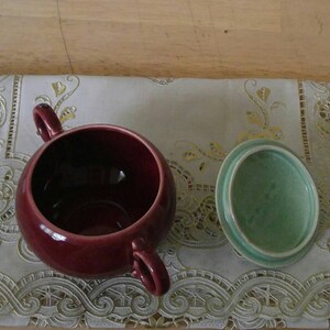 May include: A burgundy ceramic sugar bowl with a matching lid. The lid is a light green color. The bowl and lid are sitting on a white lace tablecloth.