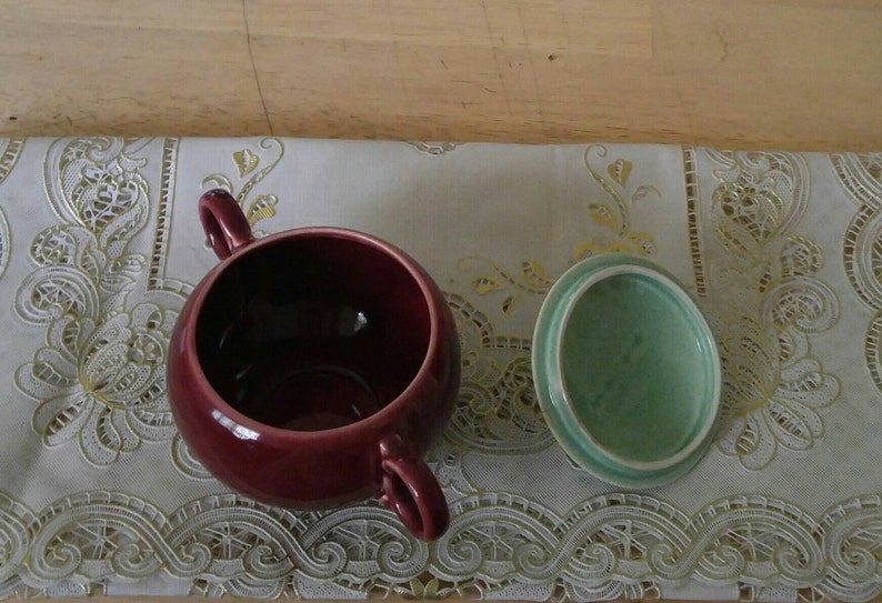 May include: A burgundy ceramic sugar bowl with a matching lid. The lid is a light green color. The bowl and lid are sitting on a white lace tablecloth.