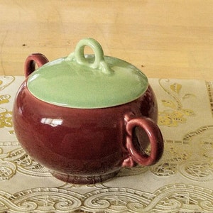 May include: A red and green ceramic sugar bowl with a lid. The bowl has two handles and a decorative knob on the lid.