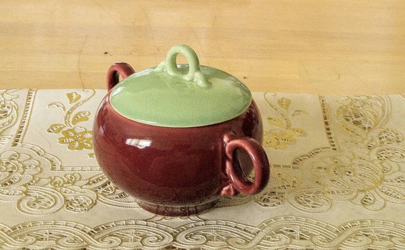 May include: A red and green ceramic sugar bowl with a lid. The bowl has two handles and a decorative knob on the lid.