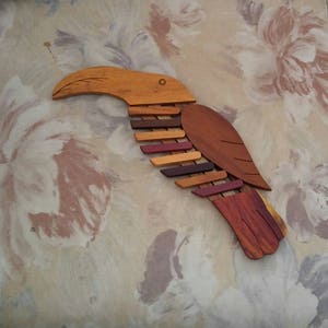 K&ouml;nnte beinhalten: Eine h&ouml;lzerne Toukan-Wandskulptur mit einem langen Schnabel und bunten Fl&uuml;geln. Der Tukan besteht aus verschiedenen Holzschattierungen, darunter Braun, Rot und Orange.