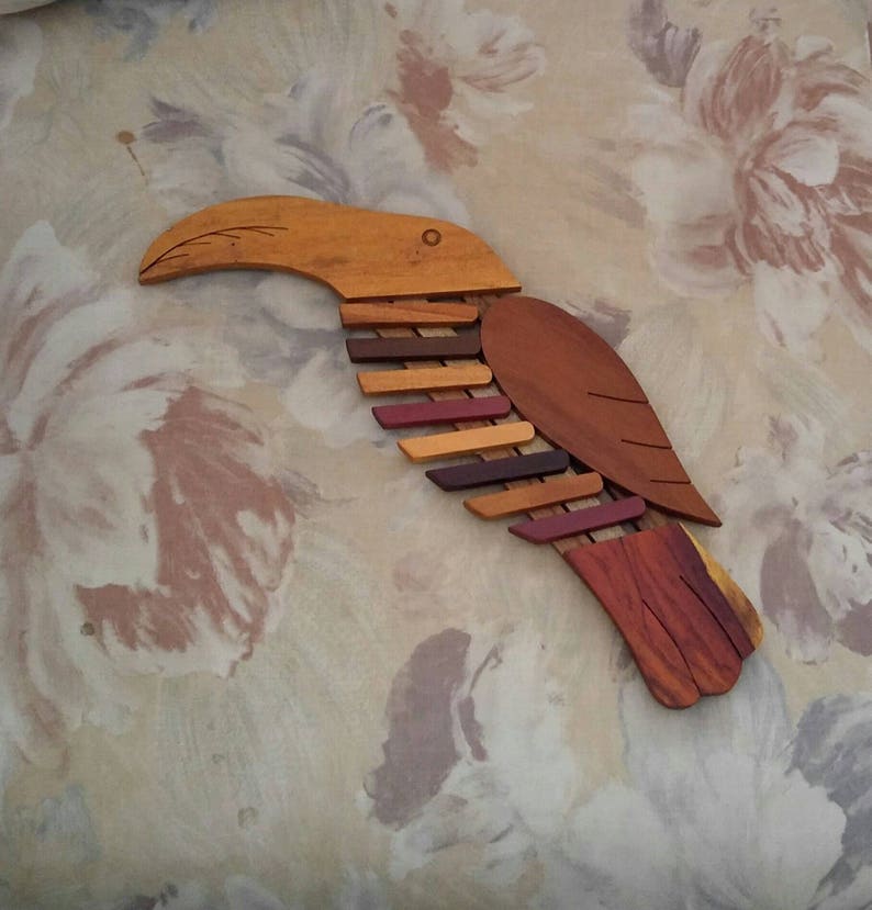 K&ouml;nnte beinhalten: Eine h&ouml;lzerne Toukan-Wandskulptur mit einem langen Schnabel und bunten Fl&uuml;geln. Der Tukan besteht aus verschiedenen Holzschattierungen, darunter Braun, Rot und Orange.