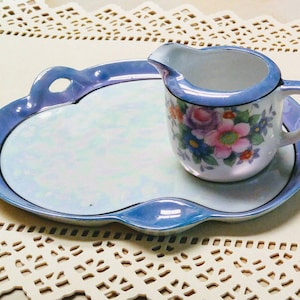 May include: A vintage porcelain creamer and serving tray set. The creamer and tray have a pearlescent finish with a blue trim. The creamer is decorated with a floral pattern in pink, purple, and blue. The tray has a unique, scalloped shape.