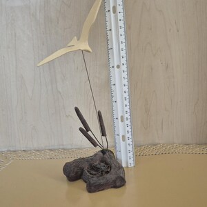 May include: A wooden bird sculpture with a long, thin neck and a wingspan of about 6 inches. The bird is perched on a small, brown wooden base with three brown reeds sticking up from the base.