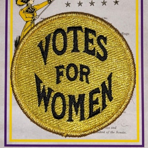 May include: A gold circular patch with black embroidered text that reads "Votes for Women". The patch has a stitched border.