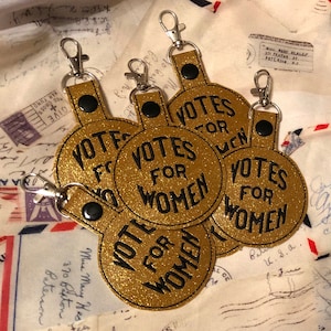 May include: Five gold glitter keychains with black text that reads "Votes for Women".