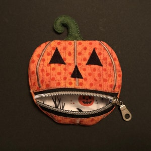 May include: A small orange pumpkin-shaped zippered pouch with a black stitched face and a green felt stem. The pouch is open, revealing a white fabric lining with a black and white pattern.