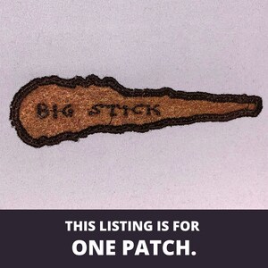 May include: Brown embroidered patch with black stitching. The patch is shaped like a long, thin stick with the words "BIG STICK" embroidered on it.