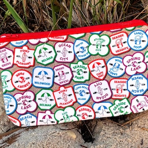 Puede incluir: Una bolsa blanca con cremallera con un patrón colorido de varios nombres de ciudades y logotipos de la costa de Nueva Jersey. La bolsa está hecha de tela y tiene una cremallera roja.