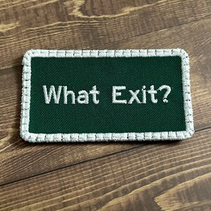 Puede incluir: Un parche verde bordado con costuras blancas alrededor de los bordes. El parche dice "What Exit?" en hilo blanco.