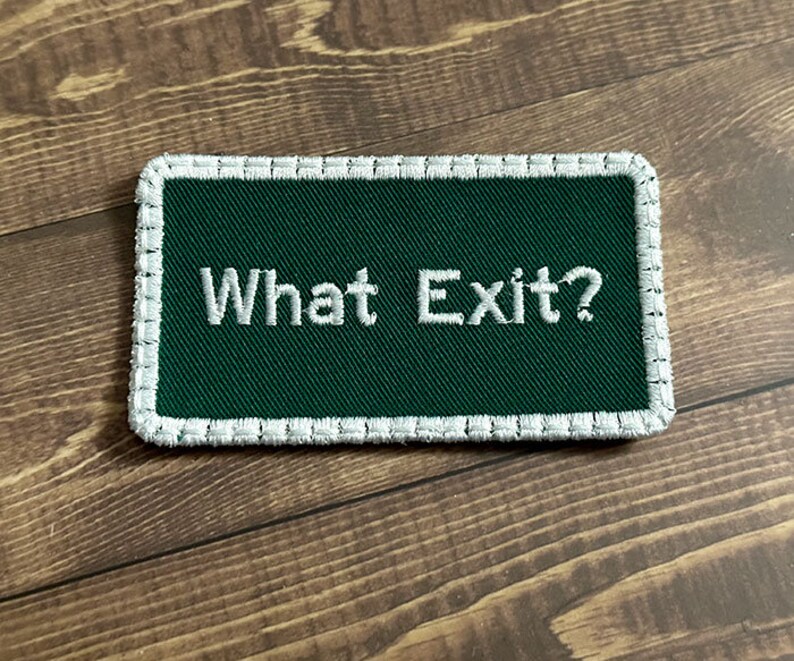 Puede incluir: Un parche verde bordado con costuras blancas alrededor de los bordes. El parche dice "What Exit?" en hilo blanco.