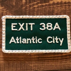 Puede incluir: Parche bordado verde y blanco con costuras blancas alrededor de los bordes. El parche dice "EXIT 38A Atlantic City".
