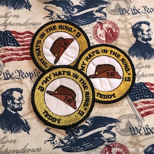 May include: Three round, gold-colored patches with black trim. Each patch features a white background with a brown and red hat, the text "TR" and the words "My Hats In The Ring '12". The bottom patch also has the word "Teddy" printed below the hat.