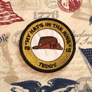 May include: A gold and black embroidered patch with the text "19 My Hats In The Ring 12" and "Teddy". The patch features a brown felt hat with the letters "TR" embroidered on the hat.