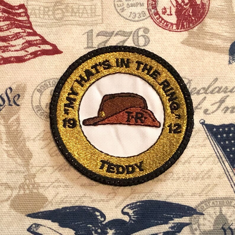 May include: A gold and black embroidered patch with the text "19 My Hats In The Ring 12" and "Teddy". The patch features a brown felt hat with the letters "TR" embroidered on the hat.