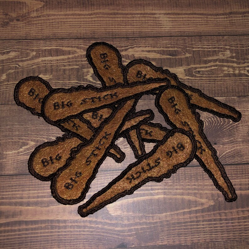May include: A pile of brown felt patches with the text "Big Stick" embroidered on them. The patches are shaped like a long, thin stick.