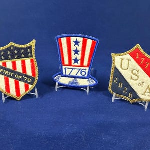 May include: Three patriotic embroidered patches on clear stands. The patches include a shield with "Spirit of '76", a top hat with "1776", and a shield with "USA of A 2026". Each patch features red, white, and blue colors.
