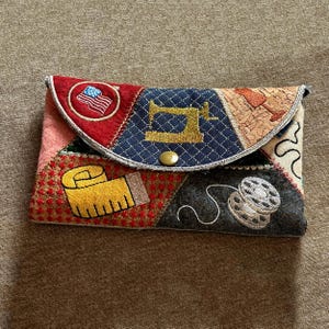 以下が含まれることがあります： パッチワークのクラッチバッグ。縫製をテーマにした刺繍が施されています。デザインには、アメリカ国旗のある赤い部分、金色のミシンがある青い部分、黄色のメジャーが含まれています。クラッチバッグは金のボタンで留められています。