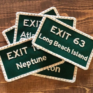 May include: Five green and white embroidered patches with the text "EXIT" and different locations. The patches are on a wooden surface.