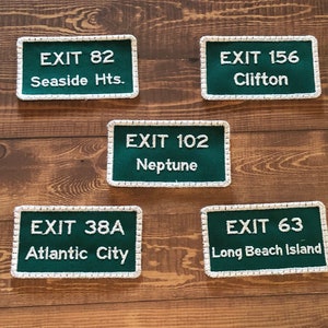 Puede incluir: Cinco parches rectangulares verdes y blancos con costuras blancas alrededor de los bordes. Cada parche tiene texto blanco que dice "EXIT" seguido de un n&uacute;mero y un nombre de lugar. Los parches est&aacute;n sobre una superficie de madera.
