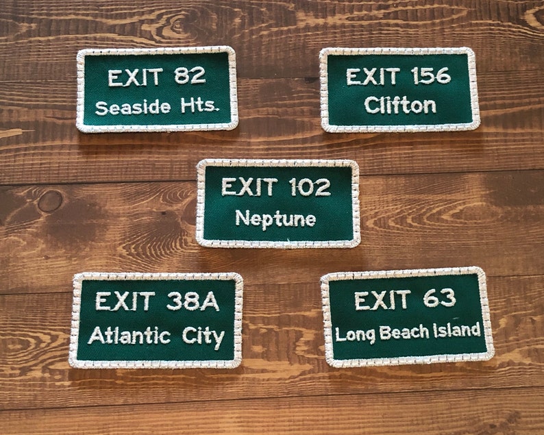 Puede incluir: Cinco parches rectangulares verdes y blancos con costuras blancas alrededor de los bordes. Cada parche tiene texto blanco que dice "EXIT" seguido de un n&uacute;mero y un nombre de lugar. Los parches est&aacute;n sobre una superficie de madera.