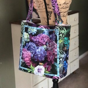 May include: A black tote bag with a floral print featuring purple and blue hydrangeas. The bag has a blue and green border around the floral print and a black and purple patterned background.