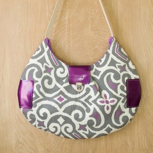 Könnte beinhalten: Eine graue und violette gemusterte Handtasche mit einer violetten Klappe und einem silbernen Verschluss. Die Tasche hängt an einem Haken an einer Holzwand.