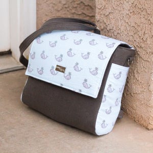 May include: A brown and blue whale-patterned diaper bag with a brown flap and a zipper closure. The bag has a strap and a handle.