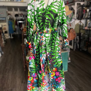 Puede incluir: Un vestido largo vintage con un vibrante estampado floral y de hojas. El vestido presenta un fondo blanco con diseños florales verdes, negros y coloridos. Tiene mangas largas, cintura con cinturón y cuello ancho. El vestido se exhibe en un maniquí.