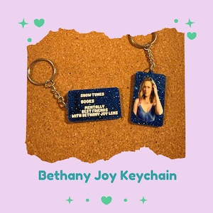 May include: Two keychains with blue glitter backgrounds. One keychain has the text "SHOW TUNES BOOKS MENTALLY BEST FRIENDS WITH BETHANY JOY LENZ". The other keychain has a photo of a woman.