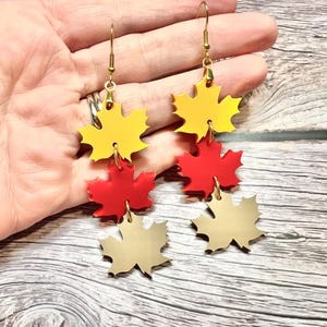 May include: Gold-colored dangle earrings featuring a maple leaf design in yellow, red, and gold. The earrings have a gold-tone hook and are made of a shiny material. The earrings are a fall-themed accessory.