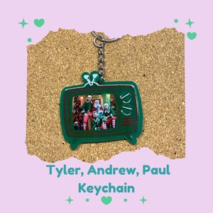 May include: Green keychain in the shape of a retro television. The television screen displays a photo of people dressed as elves with Santa Claus. The keychain has the text "Tyler, Andrew, Paul Keychain".