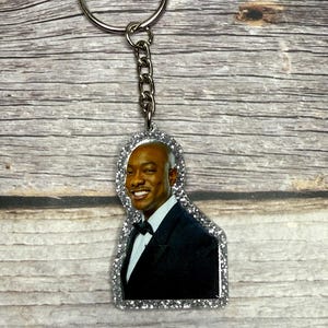 May include: A silver glitter keychain featuring a smiling man in a black tuxedo and bow tie. The keychain has a silver chain and ring. The image is set against a wooden background.