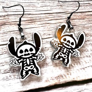 May include: A pair of black skeleton Stitch earrings. The earrings are made of clear acrylic with a black outline and a skeleton design. The earrings are hanging from black hooks.