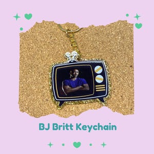 May include: A gold-colored keychain featuring a black and gold glitter-framed television with a photo of a person on the screen. The keychain has a gold chain and a small decorative top. The text "BJ Britt Keychain" is printed below the keychain.