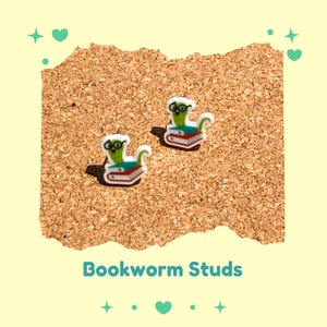 Peut inclure: Deux paires de boucles d'oreilles à clous représentant des vers de bibliothèque. Chaque boucle d'oreille montre un ver vert portant des lunettes, assis sur une pile de livres rouges et verts. Le texte "Bookworm Studs" est en dessous.