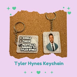 May include: Two keychains with silver key rings. One keychain has the text "SINGLE", "TAKEN", and "MENTALLY MARRIED TO TYLER HYNES" with a checkmark. The other keychain has a photo of a man in a suit. The text "Tyler Hynes Keychain" is at the bottom.
